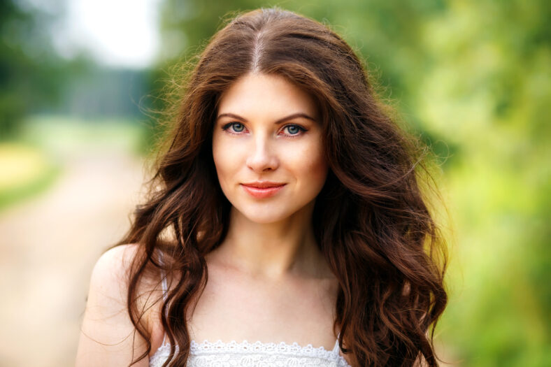 Beautiful young woman close-up in white dress, against green of park.