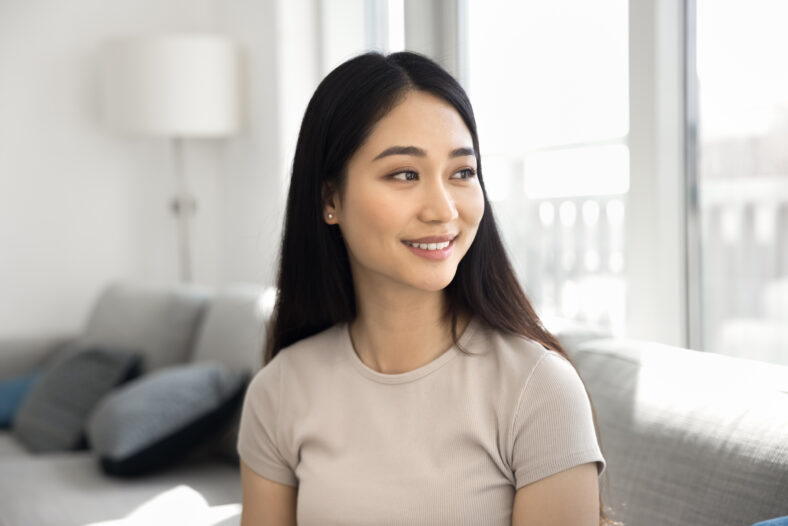 Close up shot pretty woman sit on couch in sunny living room, gazing to side with smile, looks contented, spend carefree pastime alone at own or rented apartment. Modern lifestyle, relaxation, comfort