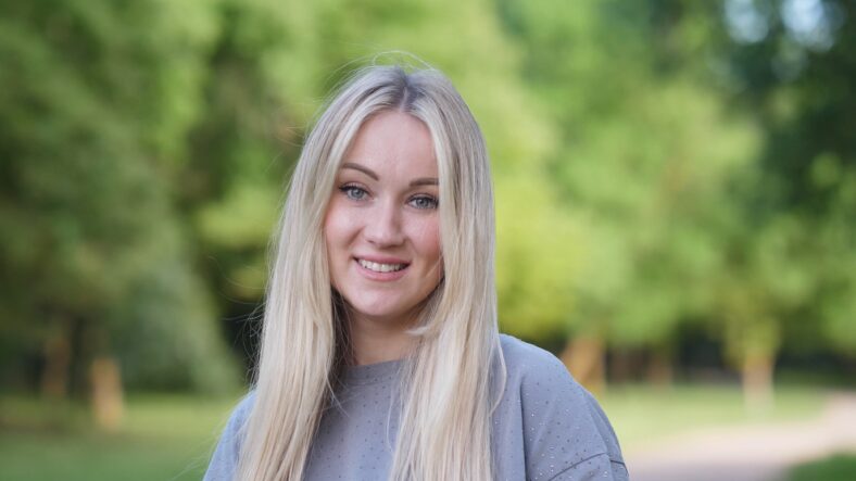 A cheerful young woman with long, flowing blonde hair beams with pure happiness in a vibrant park outdoors