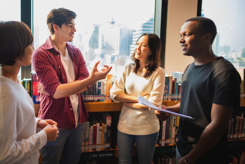 Group of diverse young adults in a library, discussing and smiling. Casual attire, diverse ethnicities, engaging in conversation, standing by bookshelves. Diverse high school students talking.