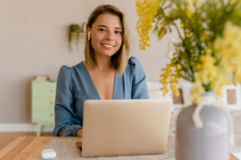 young pretty stylish woman working remote at home at table workplace, student education online, fashion blog trend, wearing blouse, smiling looking in camera, using earpods