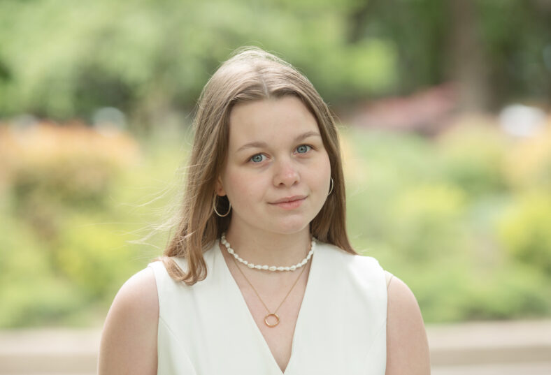young woman wearing a white dress and a pearl necklace is standing in a park. She is looking at the camera with a smile on her face