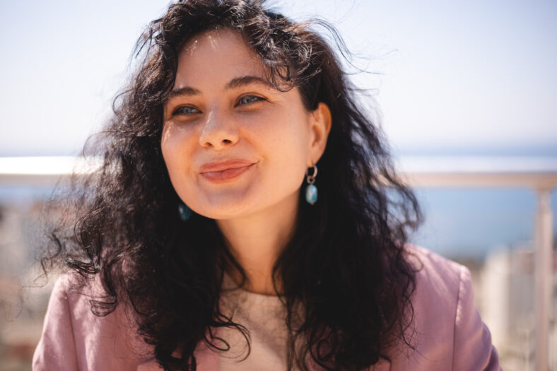 Charming brunette curly woman sitting on terrace near house, look at side, smiling, close up portrait. Girl wear pink jacket.