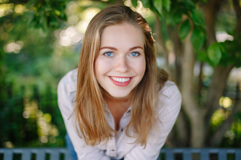 Closeup portrait of beautiful smiling white Caucasian girl woman with long blonde hair and blue eyes, wearing white shirt and jeans, outside in summer park in green foliage trees, looking in camera.