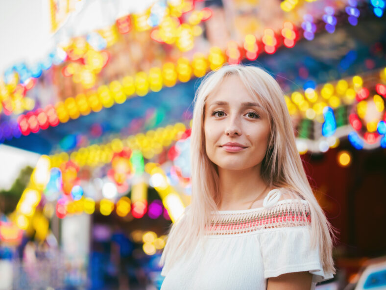 Young attractive blonde girl standing near carousel in amusement park. Beautiful portrait of woman with long hair.