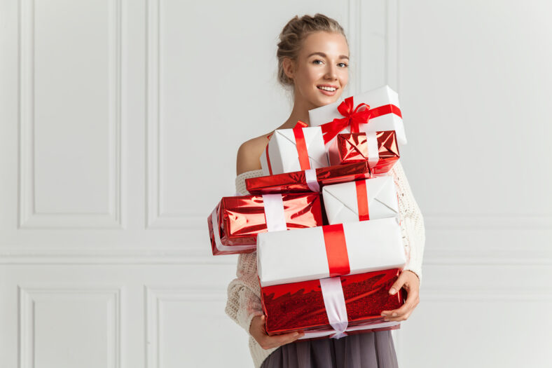 Blond hair woman in white sweater holding a lot of presents over white wall background indoor. Smiling woman holding paper gift boxes red color and looks at camera. Toothy smiling woman's face.