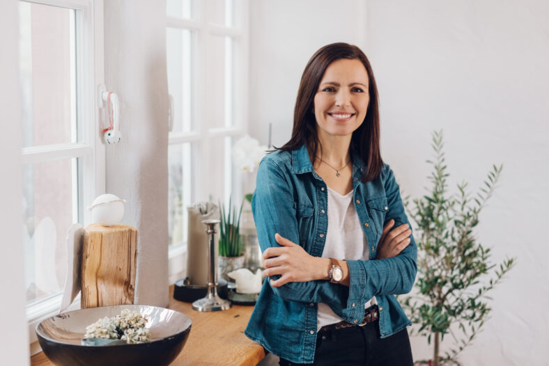 Relaxed smiling friendly woman with folded arms sitting perched on the edge of a table in her living room