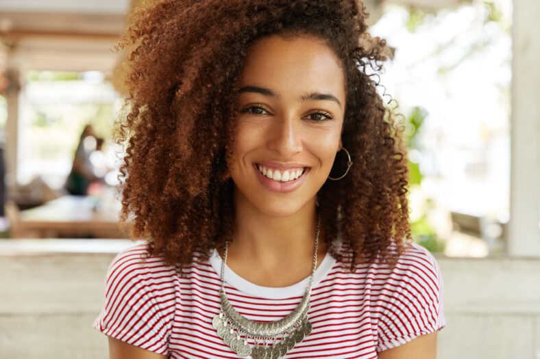 Close up shot of pretty African American female demonstrates white perfect teeth as smiles broadly, being in good mood after promotion at work, comes at cafe to celebrate this special event.