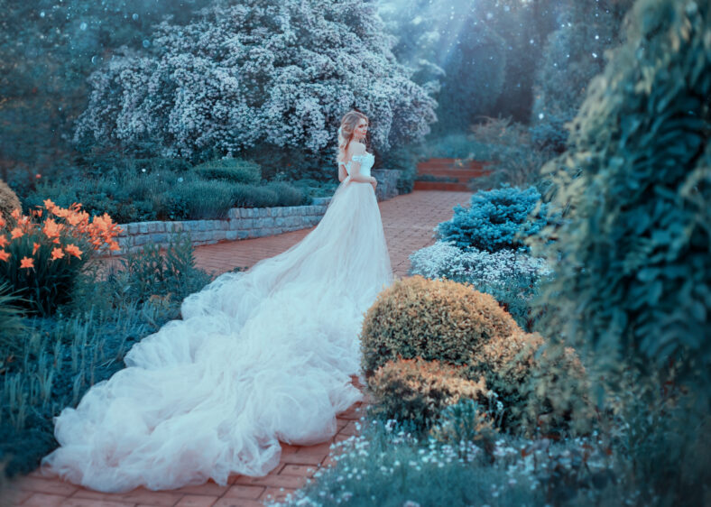 Blonde, with a beautiful elegant hairdo, walks in a fabulous blooming garden. Princess in a luxurious light pink dress with an incredibly long train goes to the next. Photo from the back. Art photo.
