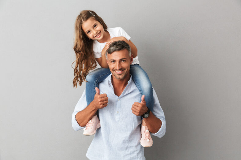 Portrait of adorable family, daughter smiling and sitting on the neck of her father 30s isolated over gray background