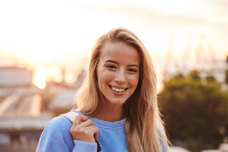Portrait of a smiling young girl with backpack standing outdoors during sunset