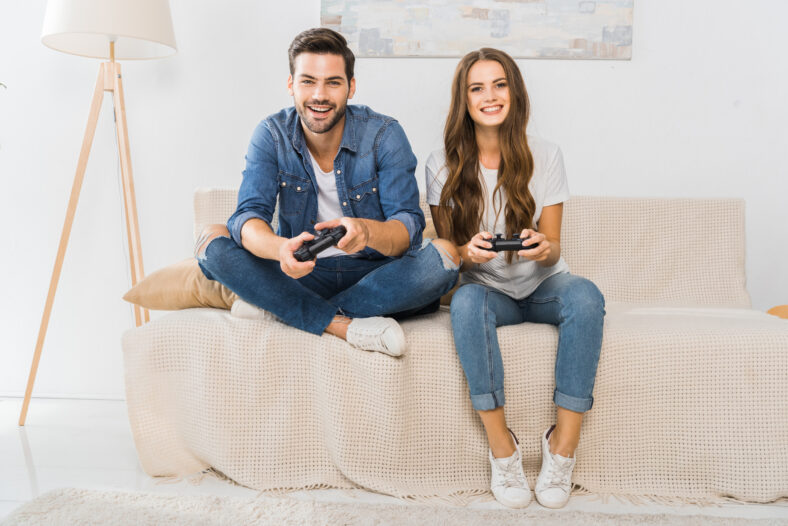 smiling young couple playing video game by joysticks on sofa at home
