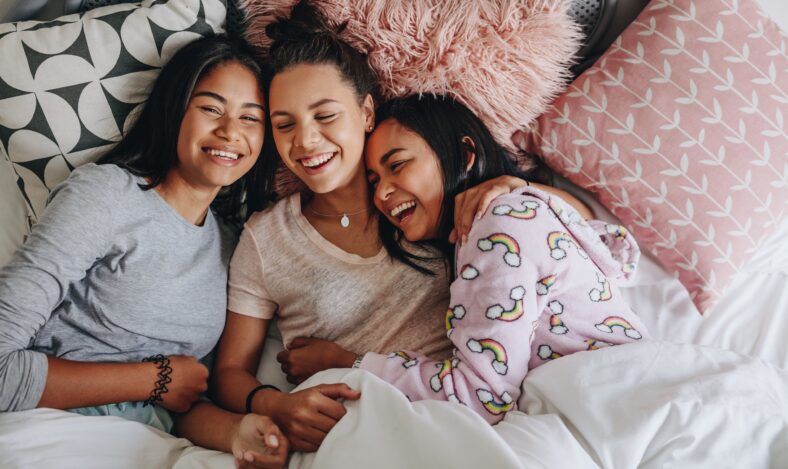 Three girls laughing and having fun while lying on bed together during a sleepover. Top view of young girls having a sleepover lying on bed and laughing.