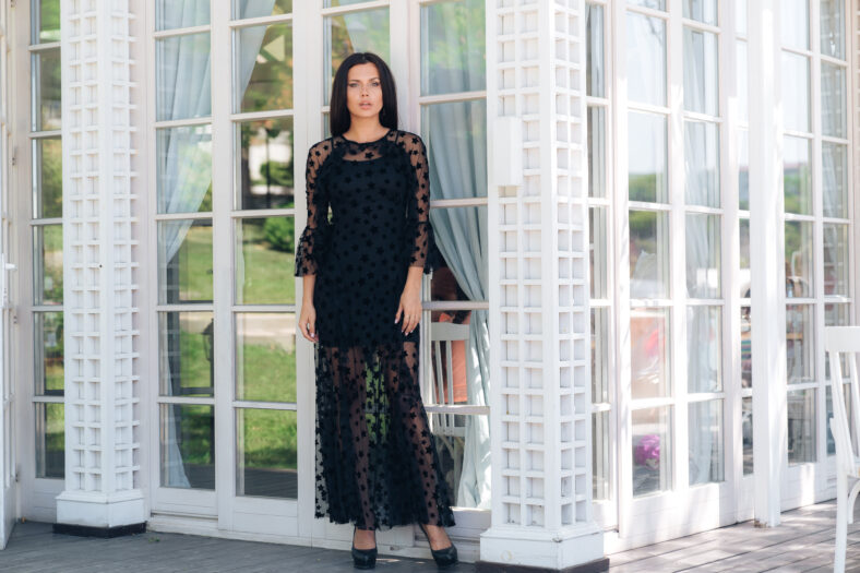 A tall slim model stands outside, dressed in a black tight-fitting mesh dress. Poses on the background of the stained-glass white doors. The concept of fashion, evening image, people, restaurants