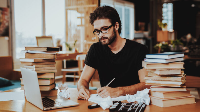 Pensive Freelance Text Writer Working at Desk. Pensive Handsome Hardworking Freelance Screenwriter Scenarist Sits Near Big Stacks of Books Writing Article, Novel, Play on Paper Sheets Looks Away