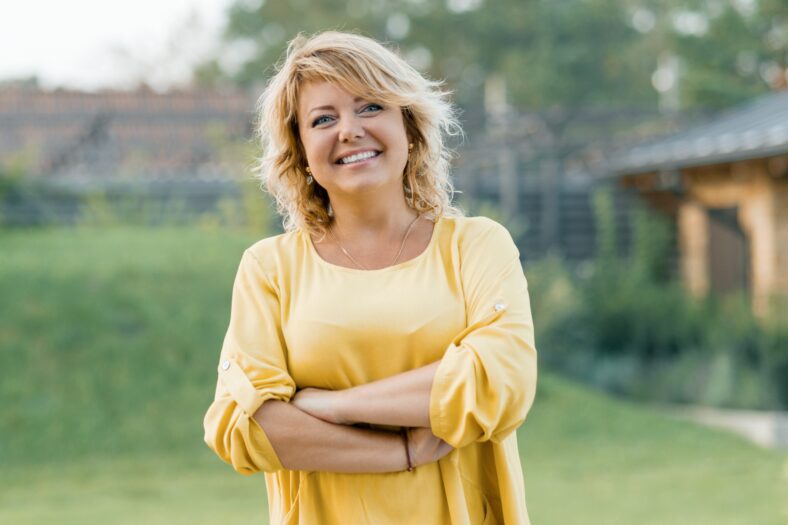 Outdoor portrait of positive confident mature woman. Smiling female blonde in a yellow dress with arms crossed near the house.