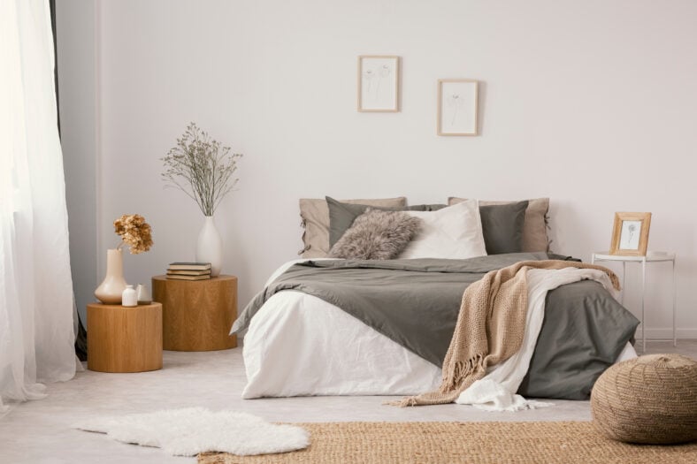 Flowers on wooden stool and pouf in white bedroom interior with posters above bed. Real photo