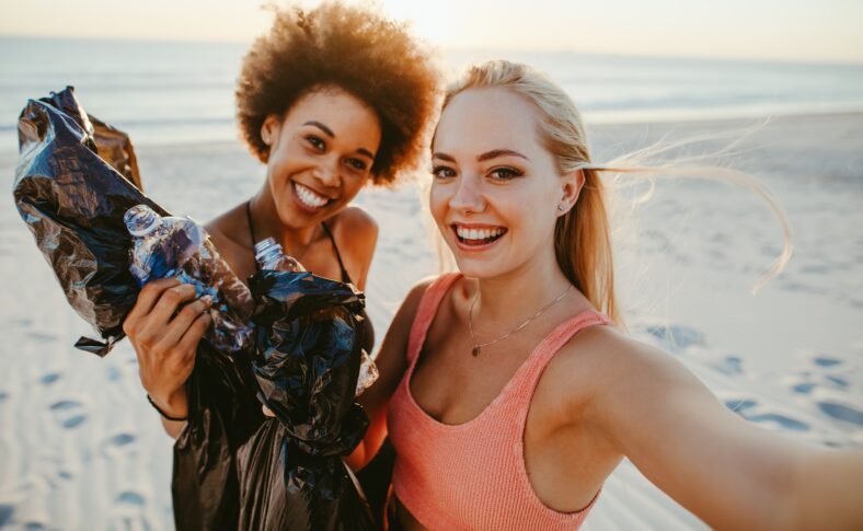Two young women holding garbage bags and taking selfie at the beach area. Two girls cleaning up rubbish on beach and taking a selfie.