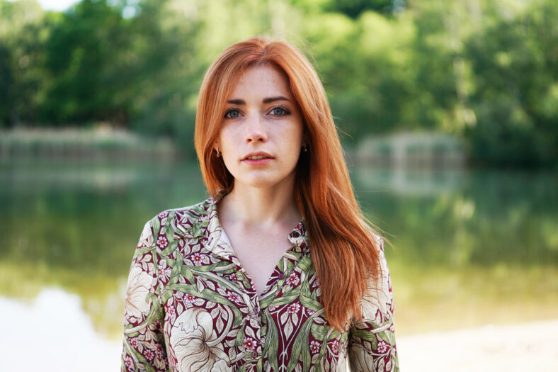 cool and confident young woman wearing floral pattern summer dress with long red hair standing by lake - authentic real people