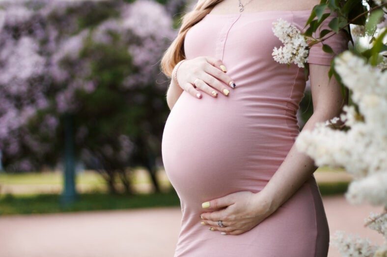 pregnant blonde girl in pink dress with bare shoulders holding hands belly with long hair on the background of lilacs and trees with a big belly on the eighth month of pregnancy close-up