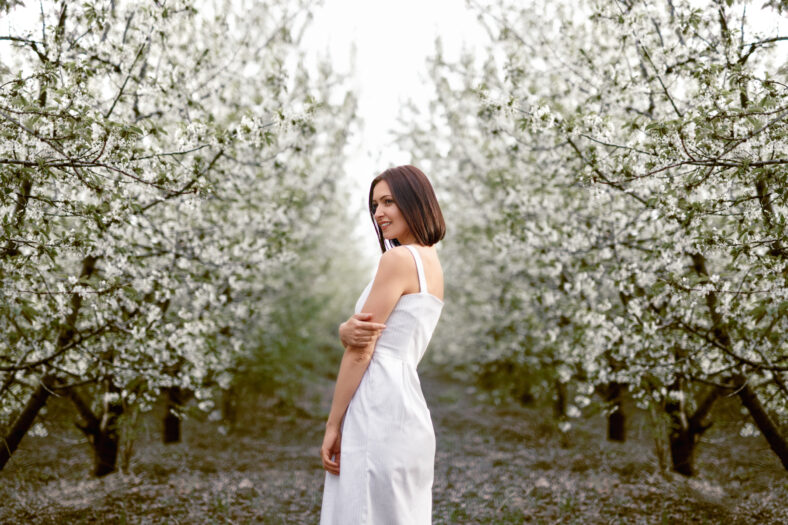 Side view of tender beautiful brunette in white dress standing among blooming spring trees smiling away