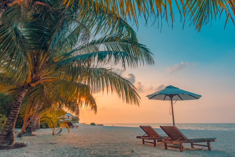 Beautiful tropical sunset scenery, two sun beds, loungers, umbrella under palm tree. White sand, sea view with horizon, colorful twilight sky, calmness and relaxation. Inspirational beach resort hotel