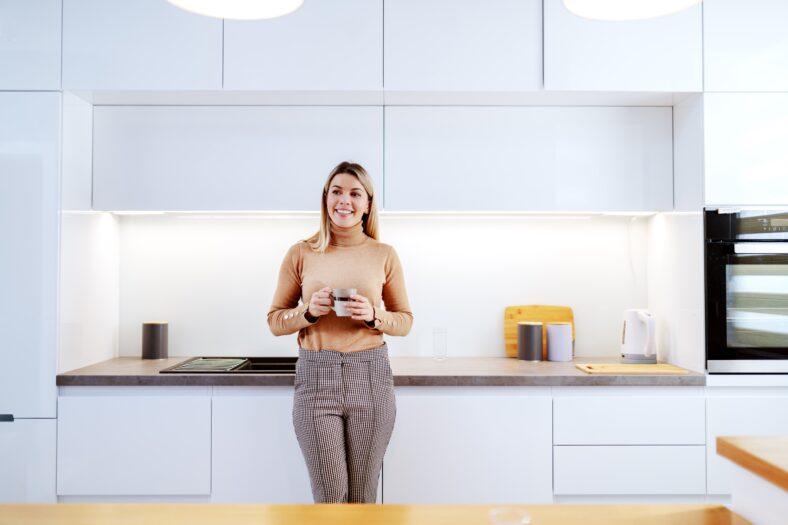 Attractive fashionable Caucasian smiling blonde woman leaning on kitchen counter and holding mug with coffee. Apartment interior.