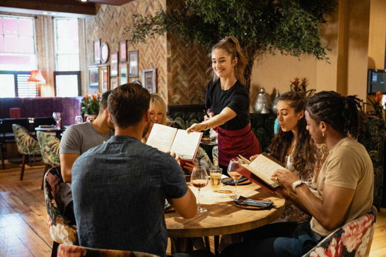 A multi-ethnic group of friends looking at menus together in a restaurant, which have been given to them by a waitress.