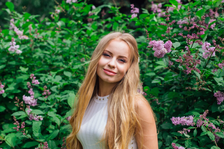 Spring portrait of a charming blond woman wearing beautiful white dress standing next to blooming purple lilac bush.