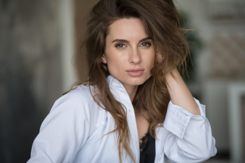 Beautiful elegant girl with flying hair and full lips in a white shirt on a gray background looks languidly at the camera. Beauty and fashionable style. Soft selective focus.