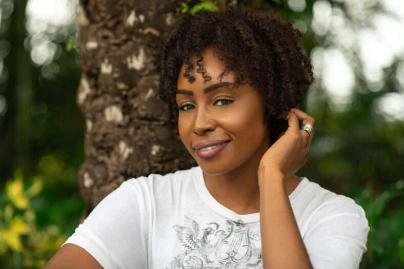 Natural portrait of an attractive black female with medium sized natural curly black & brown hair & beautiful eye makeup posing by herself underneath a palm tree.