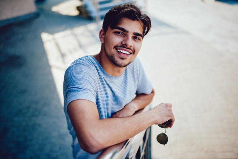 Portrait of cheerful bearded hipster guy waiting for meeting on stairs enjoying spring sunny day, happy young man with brown hair in casual outfit smiling at camera spending time outdoors