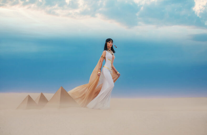young beautiful woman model image Queen Cleopatra walks in desert Egyptian pyramids. Creative retro traditional gold jewelry, bob hair cut. White satin long vintage dress, orange silk cape fly train