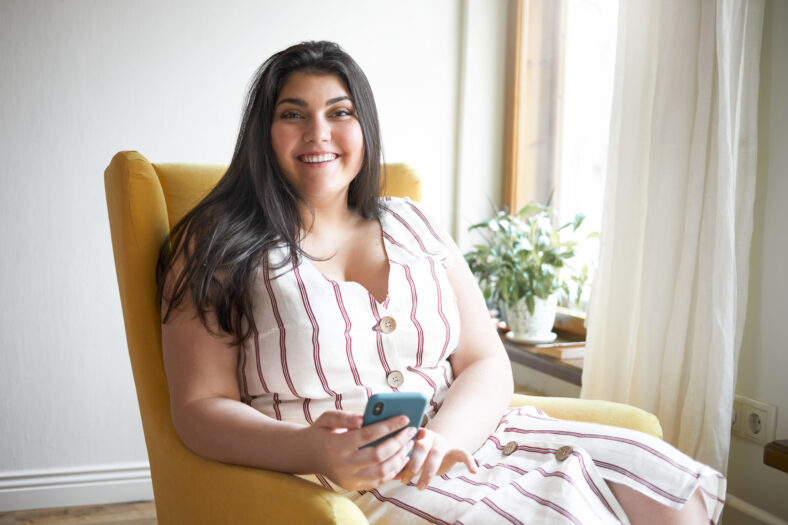 Cheerful young overweight Caucasian woman with curvy body looking at camera with broad happy smile holding smart phone, using high speed internet connection, chatting with friends online via messenger