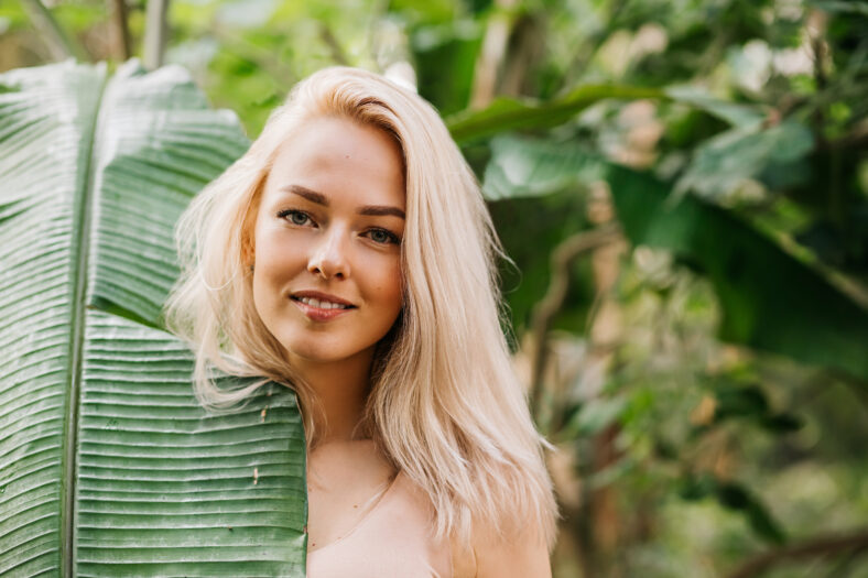 Closeup portrait of pretty caucasian lady with blonde hair, natural makeup, clean fresh skin, hiding half of face behind big tropical leaf. Skin care, spa, beauty, organic cosmetic products concept.