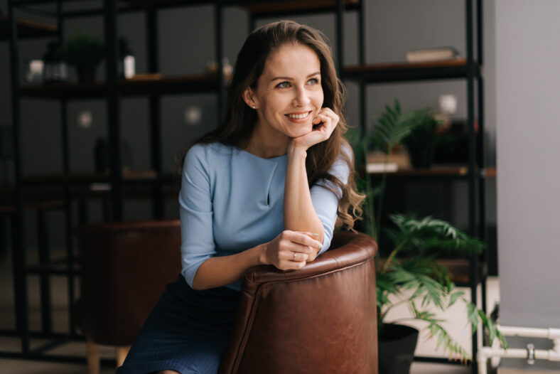 Smiling young businesswoman wearing stylish light blue dress sitting at the chair in office with modern interior, looking away. Portrait of charming business lady.