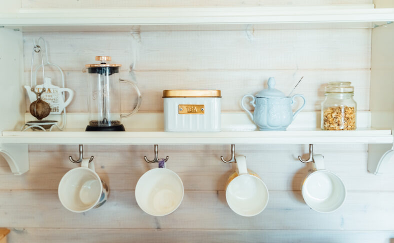 Kitchen wooden shelf with tea leaves in gold box and accessories, blue sugar bowl with spoon, strainer, press. Many colorful cups, mugs are hanging from hooks. Cozy interior in a country house.