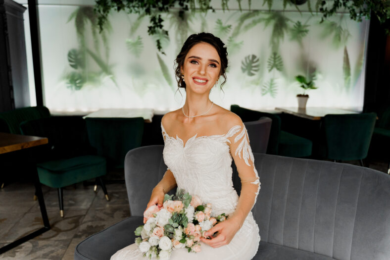Bride with wedding bouquet smiles, looks in camera and touches her face. Attractive girl portrait for social networks. Girl in wedding dress in luxury restaurant