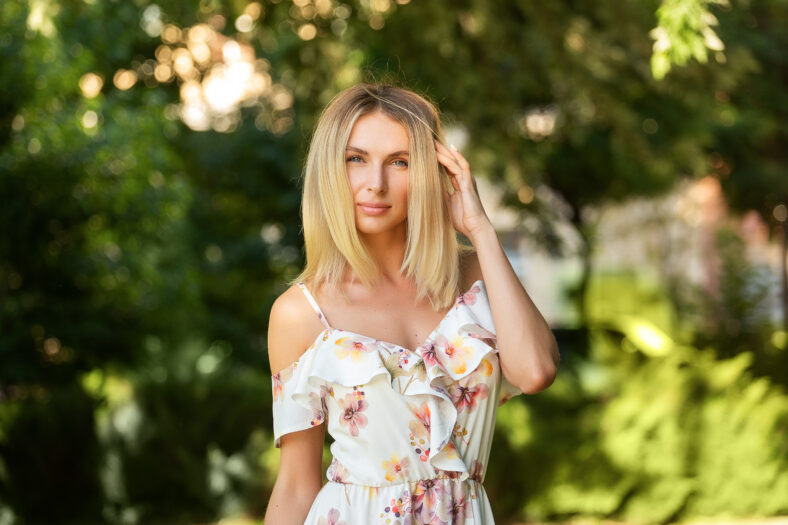 Attractive young blonde woman in a light summer dress with flowers, woman enjoying her time outdoors in a summer green park with the sunset in the background.