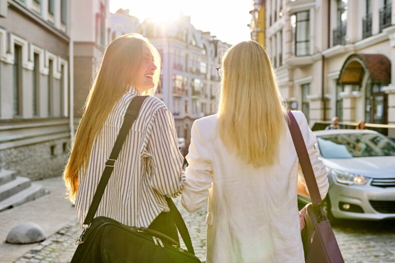 Two young women with laptop bags walking along street of sunset city, back view. Urban style background, female university students, colleagues office workers women