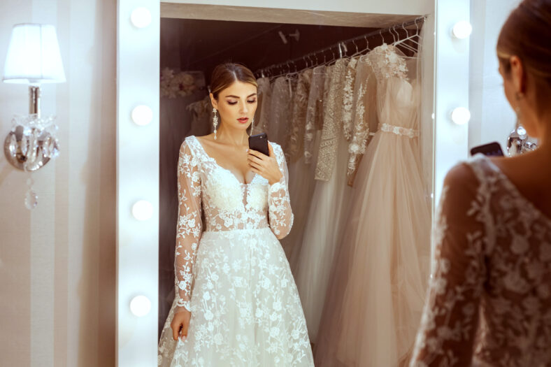 Portrait of a young bride in a wedding boutique. She takes pictures of herself in the mirror on her phone.