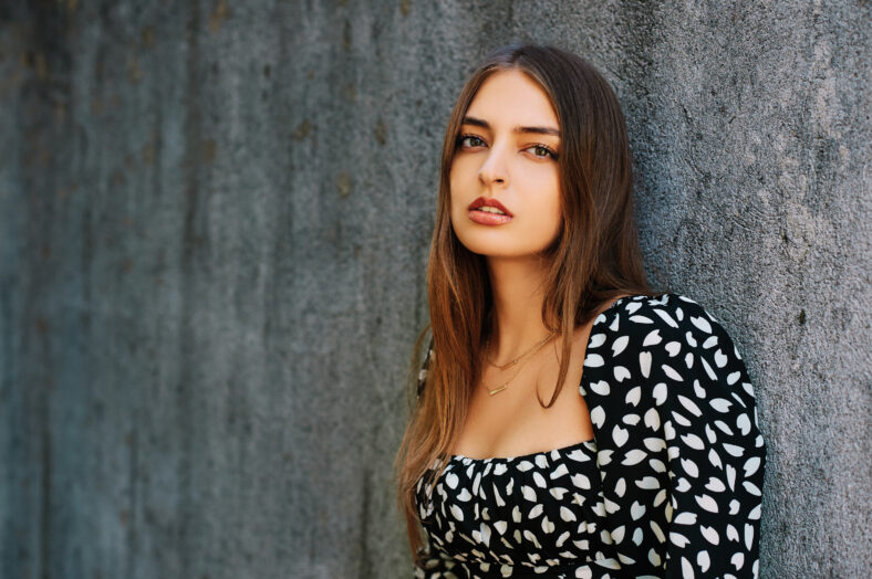 Outdoor fashion portrait of young beautiful woman wearing black and white dress, leaning on grey wall
