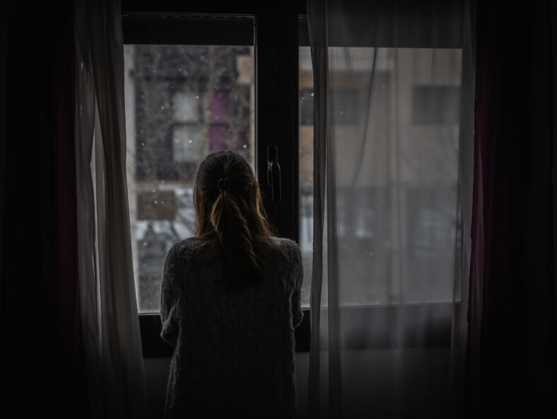 Young woman looks out a window between the curtains while it is snowing.