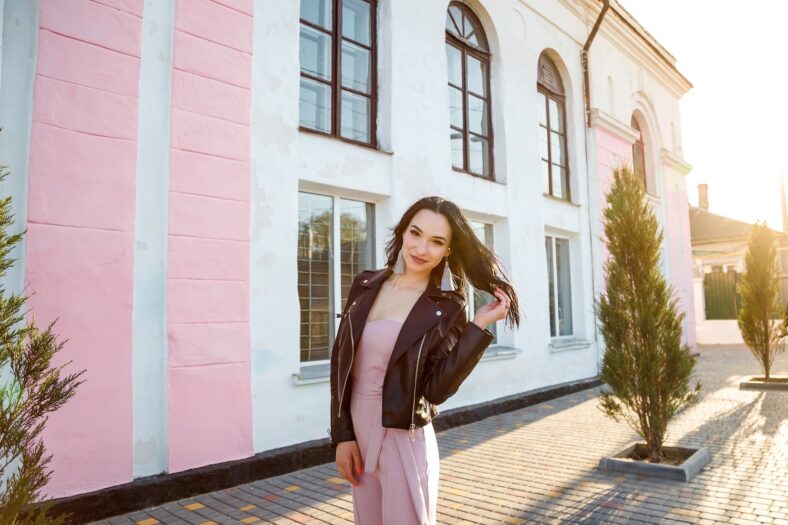 Pretty long-haired young woman in black leather jacket posing on the street by sunset light, lifestyle portrait.
