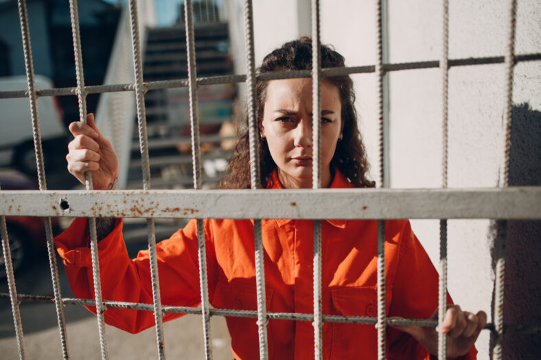 Young woman in orange suit behind jail bars. Female in colorful overalls portrait. Law and justice concept