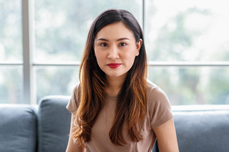 Close up portrait head shot of Asian long brown hair young beautiful female wearing t-shirt standing smiling in front of blurred green background.
