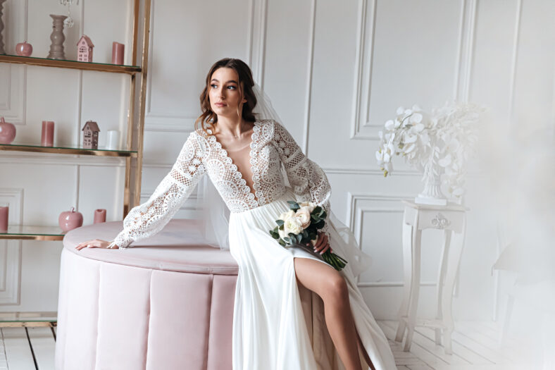 An attractive young bride in a beautiful boho wedding dress posing with a bouquet in a bright room. Wedding photo session, bride's morning.