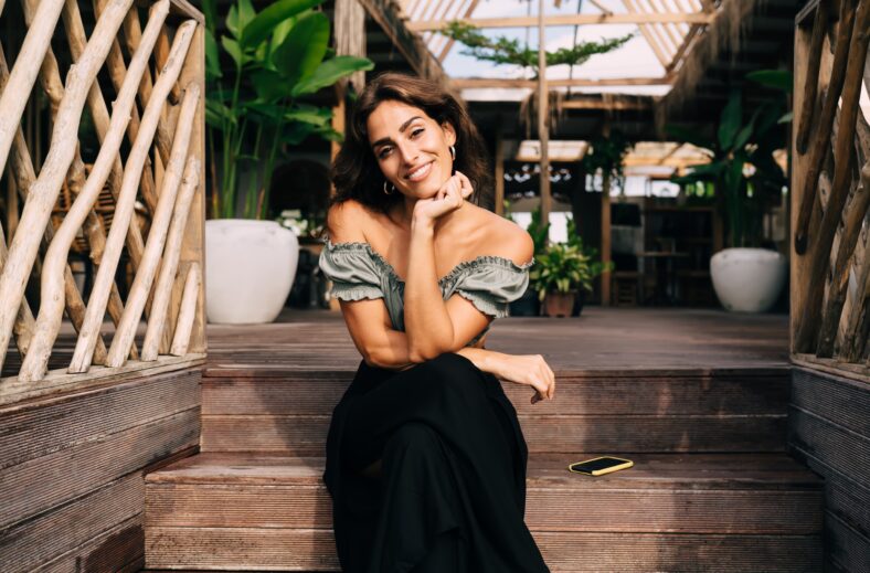 Cheerful female in trendy outfit resting on wooden steps leaning head on hand while sitting alone in sunny day against plants decorating cafe space
