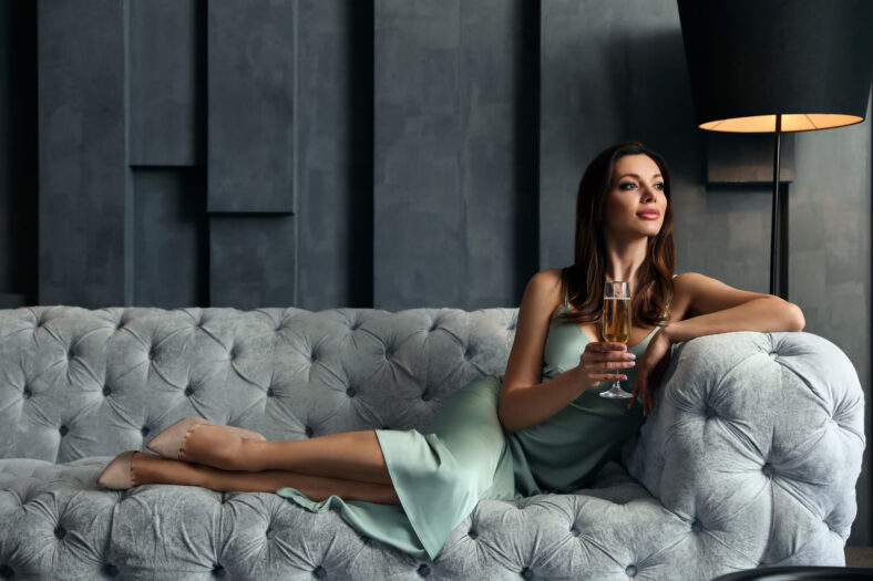 Beautiful woman in elegant dress with glass of champagne on sofa indoors. Luxury lifestyle