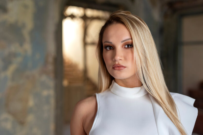 Beautiful young long haired blond woman dressed in white dress with serious face expression stand inside of rustic hall of old house